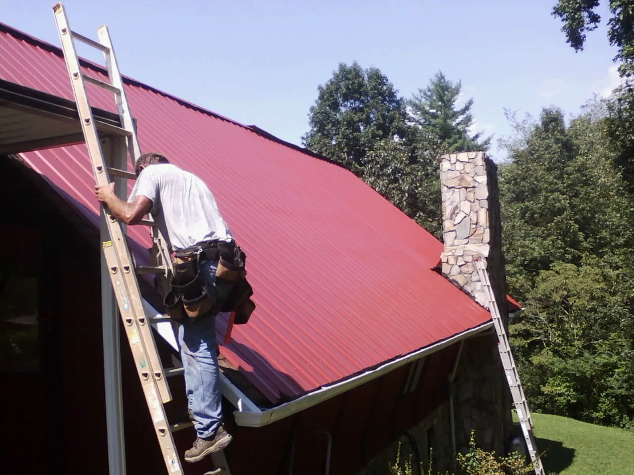 Roofer on a ladder beside a red metal roof and stone chimney — Soffit & Fascia Repair by Roofing Brothers in Pine Mountain Valley